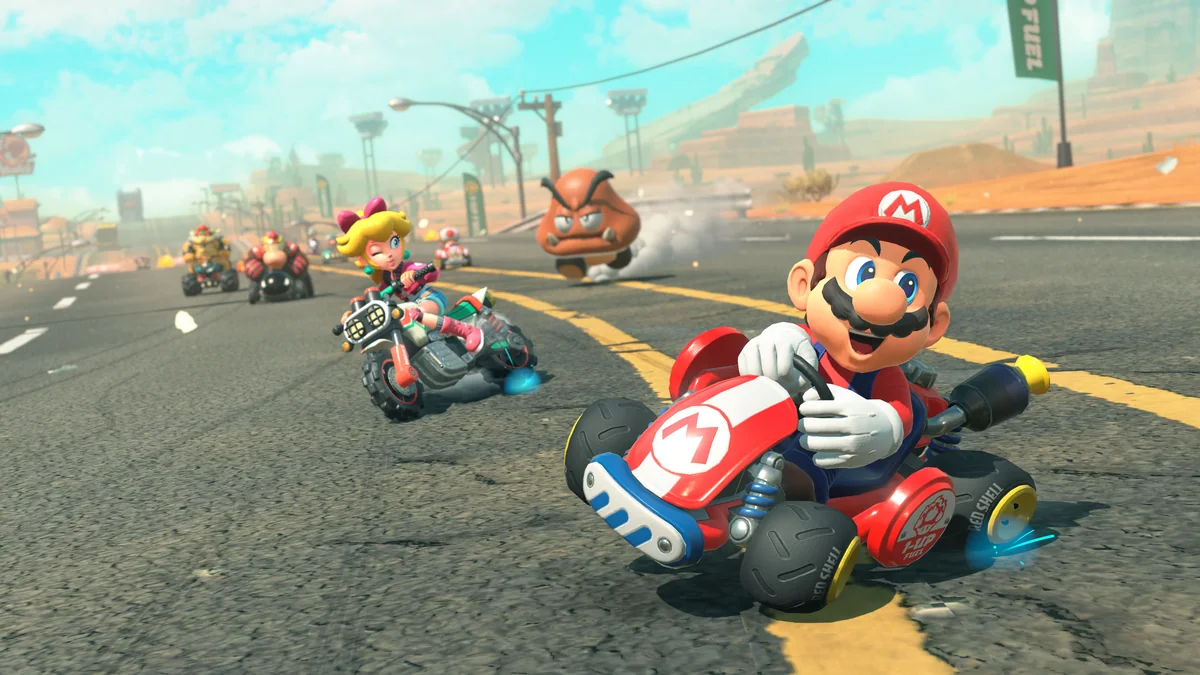 Mario drifting a red kart on a desert track, with Peach on a bike and a Goomba in the background